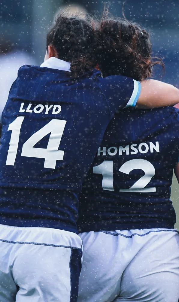 Two Scotland rugby players embrace on the rugby field. Their backs to the camera with their names and numbers printed on the back of the shirts
