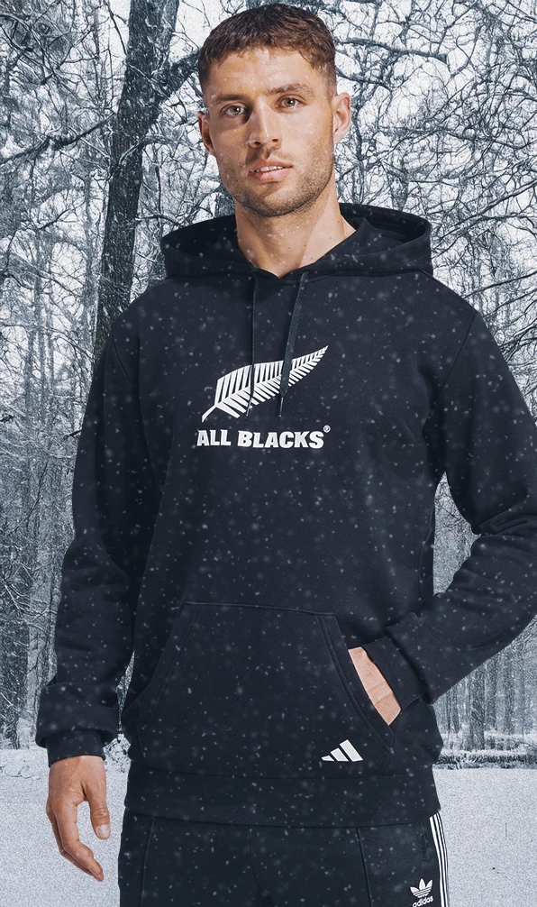 A man stands wearing a New Zealand All Blacks hoodie, in front a snowy outdoor scene while snow gently falls.