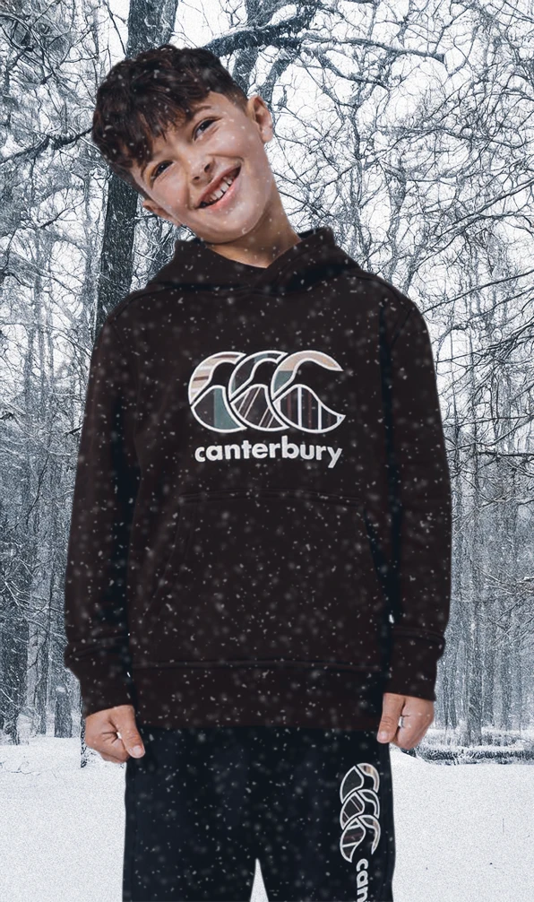 A young child wear a Canterbury of New Zealand rugby hoodie, standing in front of a snowy outdoor scene while snow gently falls