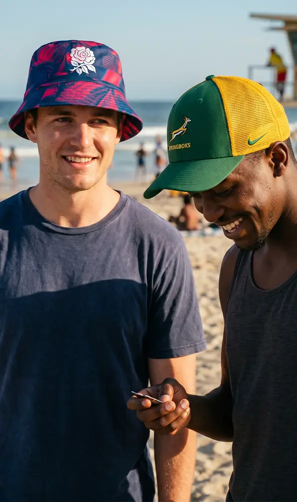 Two guys are wearing a England bucket hat and South Africa cap at the beach.