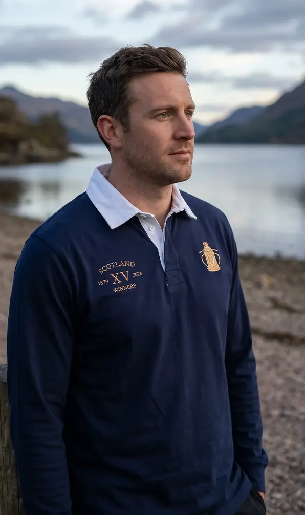 A man stands on the edge of a loch in the Scotland 2026 Cup Winners classic rugby shirt.